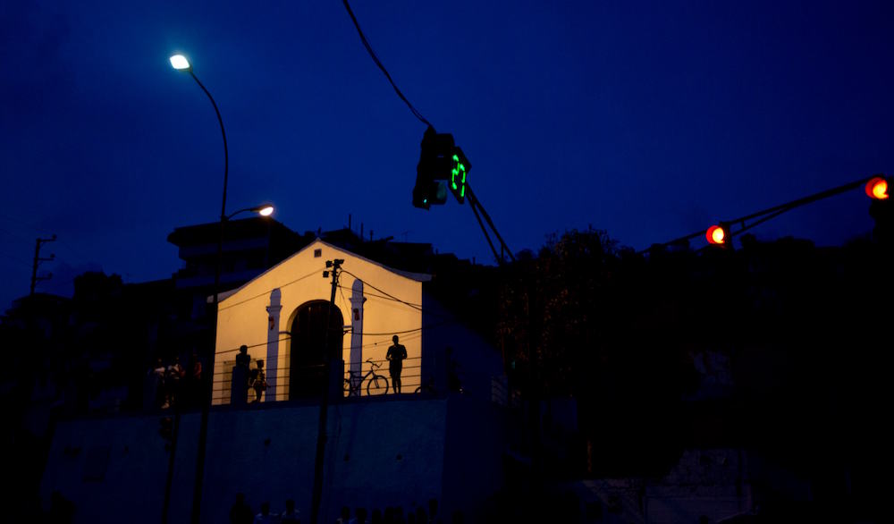 In this Saturday, April 23, 2016 photo, residents are silhouetted against the facade of a church, the only building illuminated after a 24-hour blackout, in the El Calvario neighborhood of El Hatillo, just outside of Caracas, Venezuela. Energy rationing has been added to the hardships faced by Venezuelans overwhelmed by inflation, shortages of food and medicine and rising crime. (AP Photo/Fernando Llano)