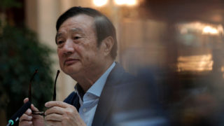 Ren Zhengfei, um senhor asiático aparentando algo em torno de 70 anos de idade. Ele veste um blazer cinza e uma camisa azul claro e segura óculos escuros fora do seu rosto. Ele está levemente virado para a esquerda.