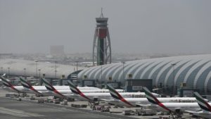 Foto do Aeroporto Internacional de Dubai. Há dois galpões e uma torre de controle ao fundo. Na frente, sete aeronaves da Fly Emirates está paradas, voltadas para os galpões.