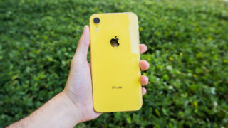 Apple iPhone XR amarelo na mão esquerda de uma pessoa. As costas do aparelho estão viradas para a câmera. Ao fundo, um gramado.