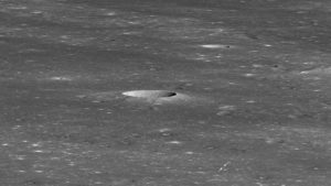 Imagem em preto e branco da superfície do lado oculto da Lua. Há uma cratera bem no centro da imagem e duas setas pequenas indicando um ponto minúsculo no canto inferior direito.