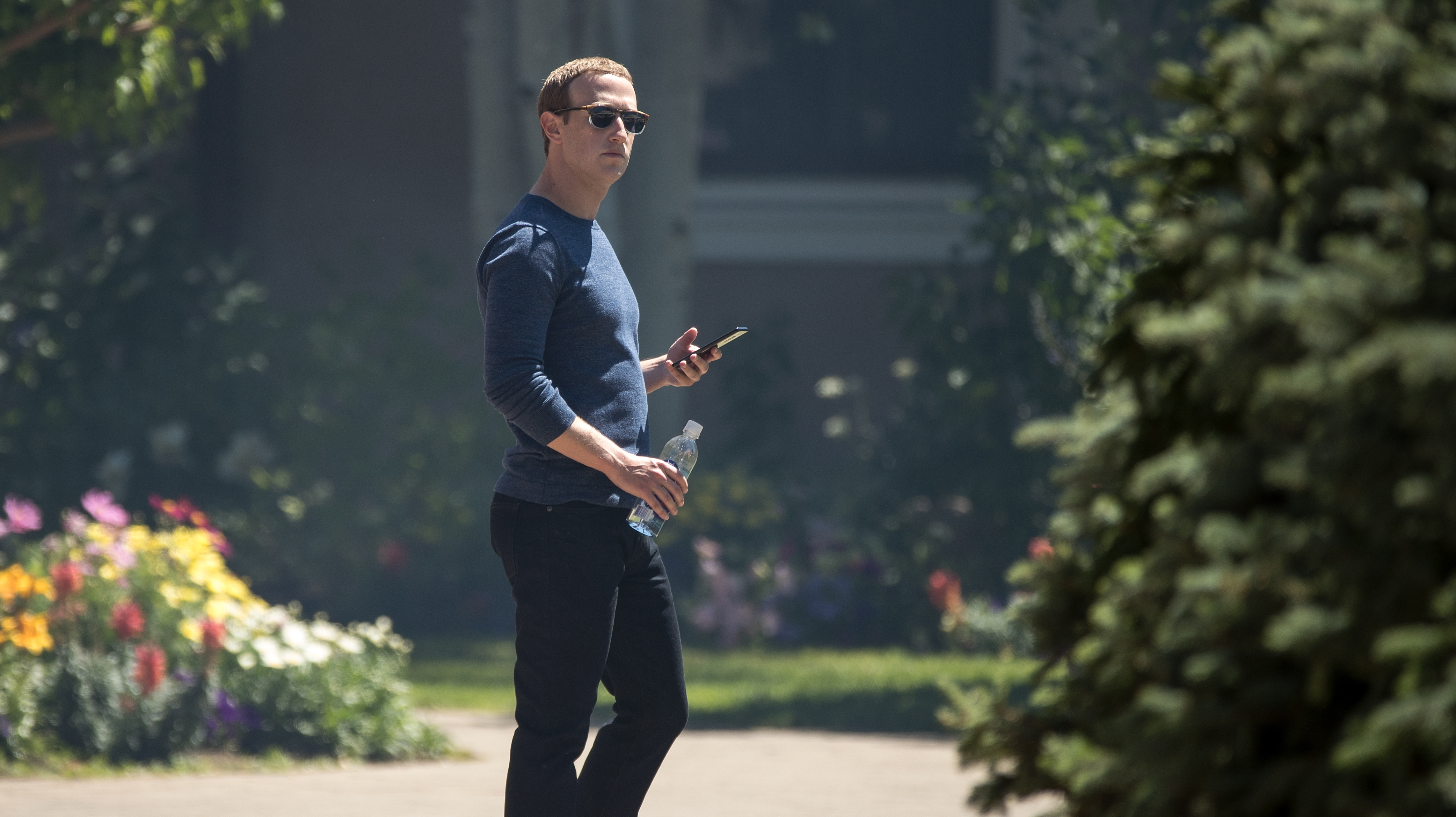 Mark Zuckerberg, CEO do Facebook, em um ambiente externo. Ele usa óculos escuros e tem uma garrafa d'água em uma das mãos e um smartphone na outra.