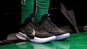 Foto dos pés do jogador de basquete Jayson Tatum. Eles estão vestindo meias verdes e calçando os tênis da Nike, que são pretos, com a sola branca, e sem cadarços. O chão da quadra também é verde, cor do Boston Celtics.