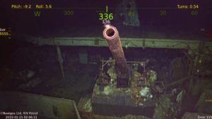 Imagem subaquática mostra um canhão de cor rosada e tamanho grande no porta-aviões, que está bastante deteriorado.