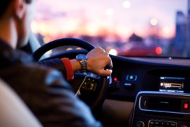 Imagem de motorista dirigindo carro. O ponto de vista é do passageiro do banco traseiro. O motorista tem uma das mãos no volante, e a outra abaixada. Pelo para-brisa, dá para ver o entardecer.