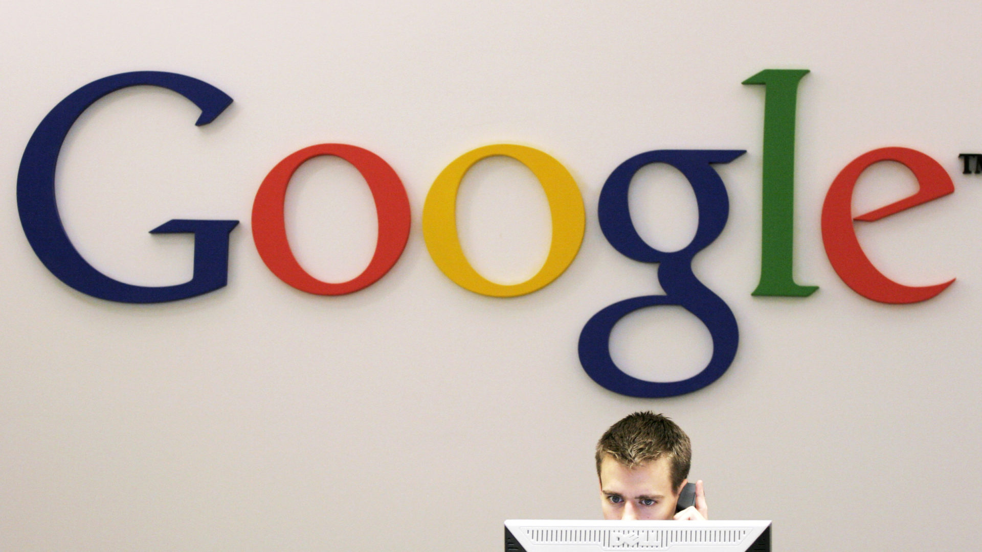 parede de escritório com o logo do Google. há um funcionário trabalhando em frente a ela. a parte superior da cabeça do funcionário está visível por trás de um monitor.