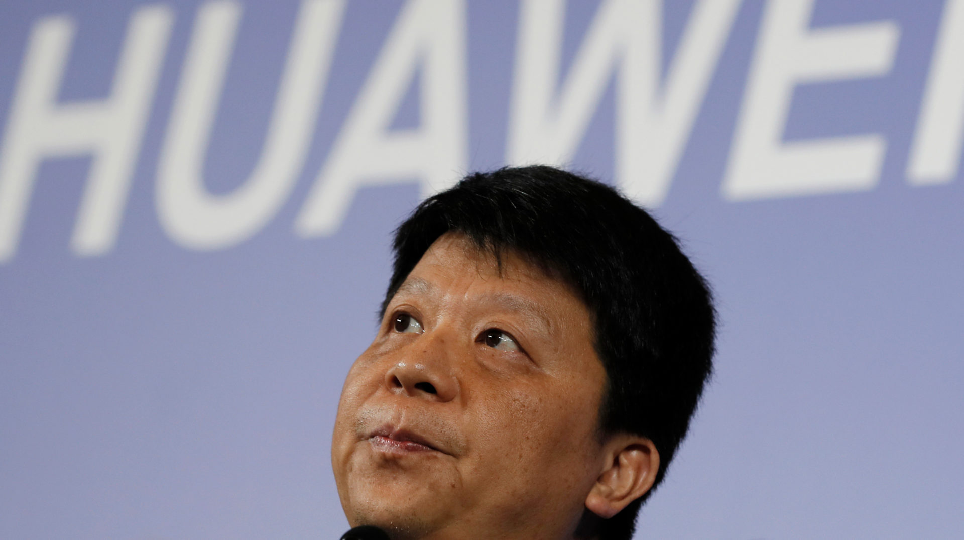presidente rotativo da Huawei, Guo Ping. Foto em close. Ao fundo, está o logo da Huawei, em branco, sobre uma projeção azul.