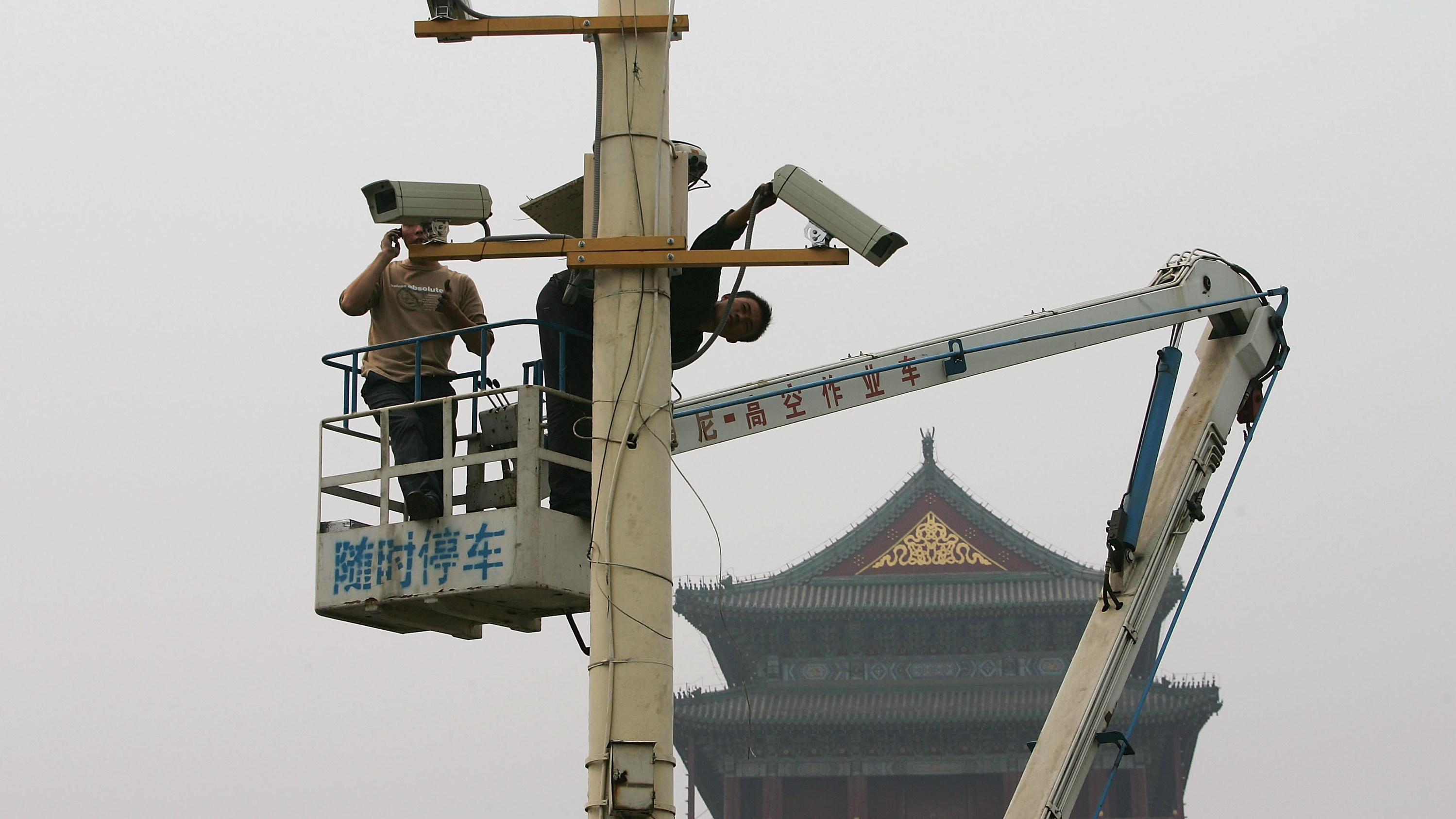 câmera sendo instalada na china. há duas pessoas em um guindaste, onde se vê logogramas chineses. ao fundo, um telhado de um templo oriental.