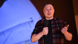 Elon Musk usando uma camisa vermelha xadrez com um microfone na mão durante uma apresentação