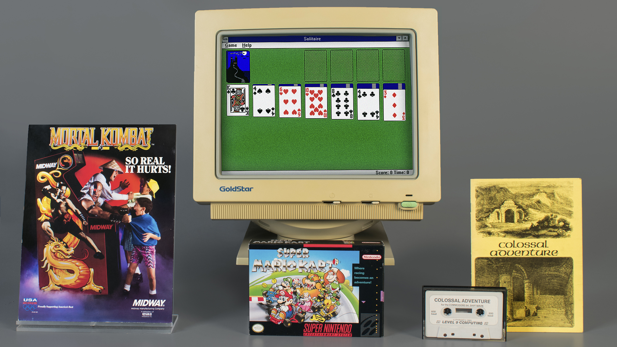 Um monitor de computador com Paciência, caixas de Mortal Kombat e Super Mario Kart, uma fita cassette e um caderno de Colossal Cave Adventure