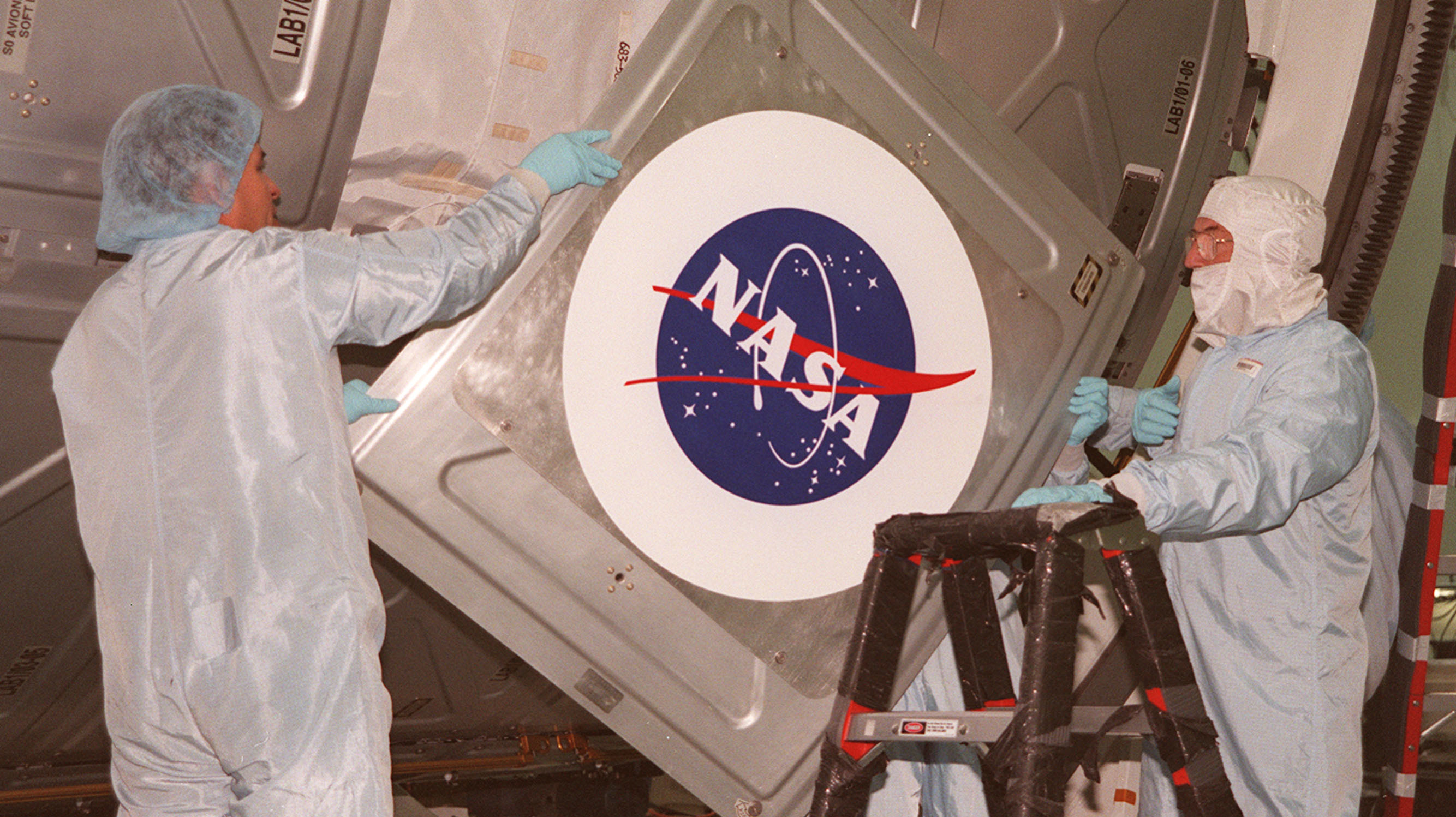 Duas pessoas, usando roupas de proteção, manuseiam uma porta de espaçonave com o logo da NASA.