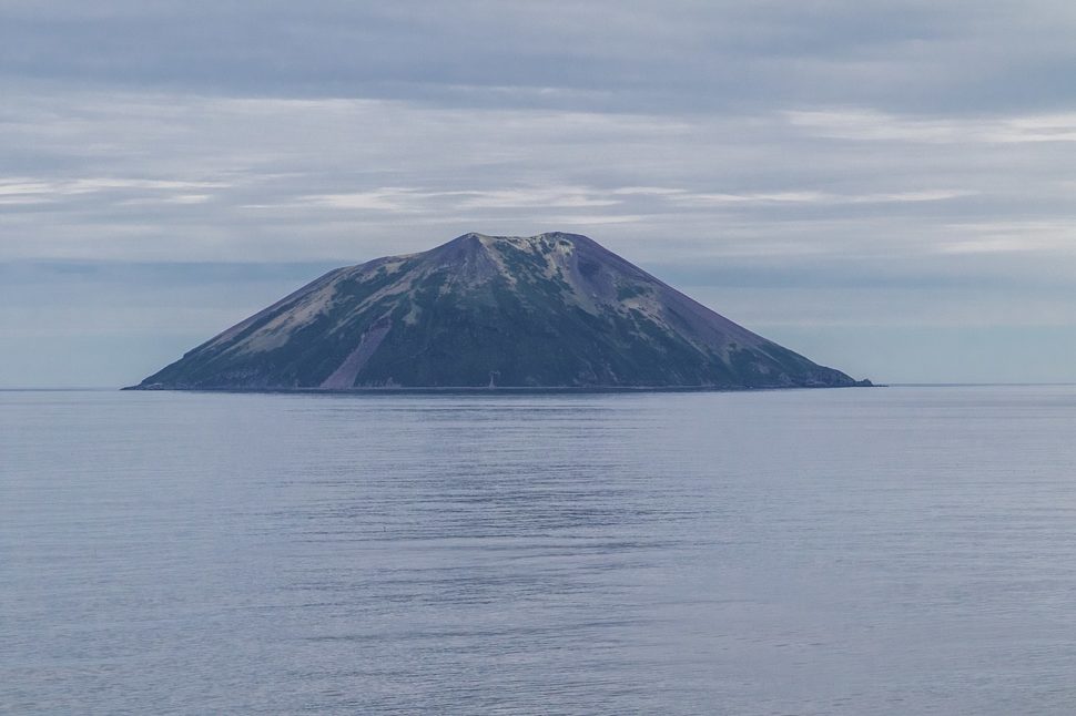 Ilha no mar. A ilha tem um formato próximo de um trapézio e está quase toda coberta por uma vegetação escura.