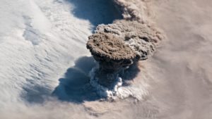 Imagem aérea da erupção do vulcão Raikoke. A metade esquerda é formada por nuvens brancas. No meio da imagem, há a pluma do vulcão se erguendo sobre as nuvens. Ela é mais escura, em um tom entre o cinza e o marrom. Na metade direita da imagem, há nuvens mais escuras, acinzentadas.
