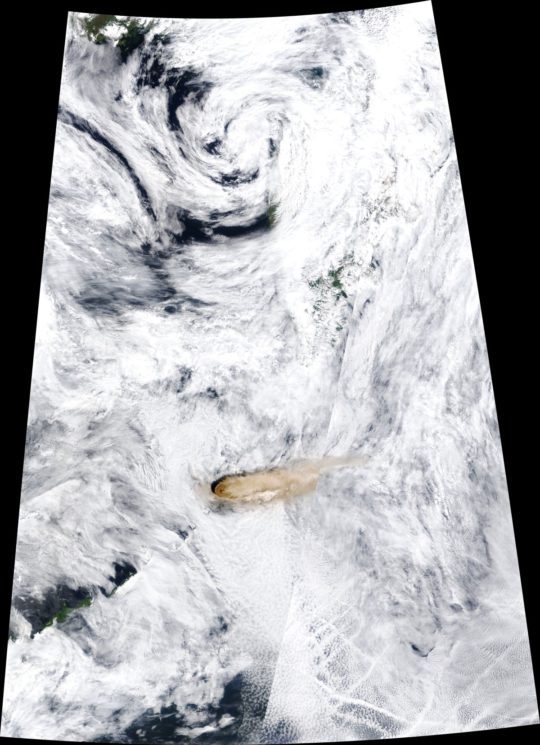 Imagem de satélite formada quase que inteiramente por nuvens brancas, com algumas falhas mostrando o azul escuro do mar por baixo delas. Pouco abaixo da metade da imagem, há um ponto em bege escuro, com nuvens em bege se espalhando para a direita.