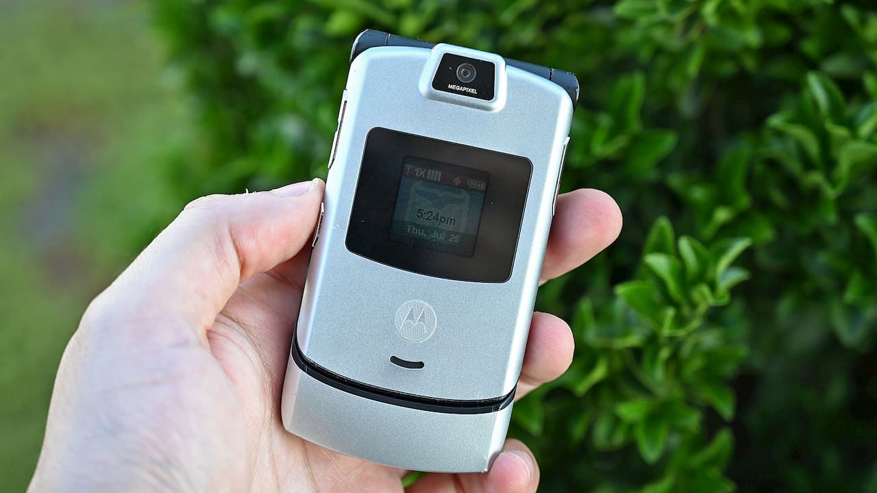 Motorola V3 clássico na cor prata sendo segurado por uma mão esquerda. O aparelho é menor que um smartphone atual, tem uma tela secundária no meio da tampa e uma câmera na parte superior.