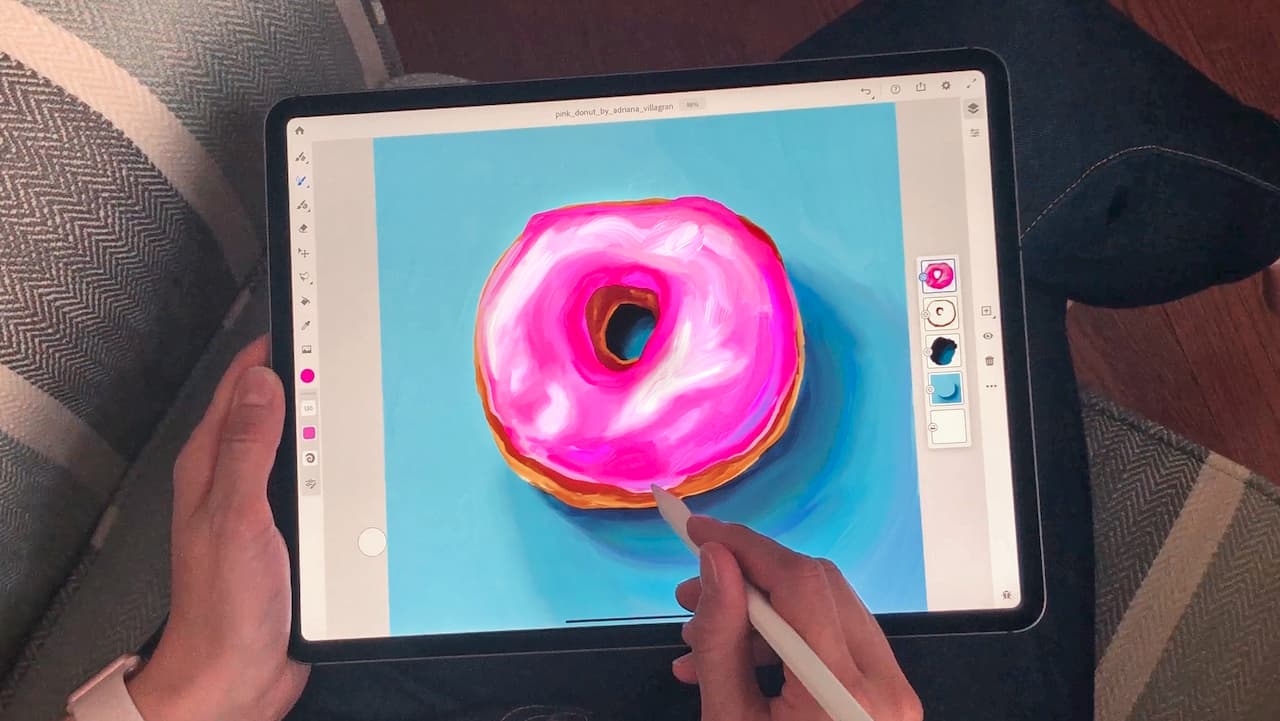Foto de uma pessoa usando o Adobe Fresco em um iPad. Há um donut cor de rosa na tela. A pessoa está segurando um Apple Pencil. Na parte de esquerda da tela, há uma coluna de ferramentas. Na parte direito, camadas com as diferentes cores usadas no donut e no fundo.