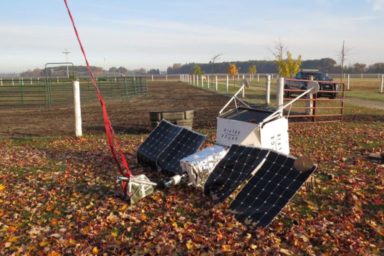 Aparelho parecido com satélite, mas com materiais claramente mais comuns e menos resistentes, caído no chão de uma fazenda.