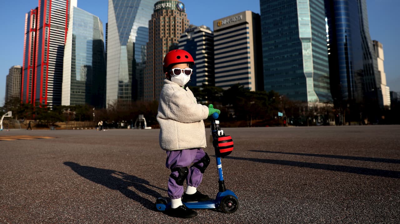 Criança de touca, óculos escuros e máscara cirúrgica. A criança está sobre um patinete. Ao fundo, vários prédios.