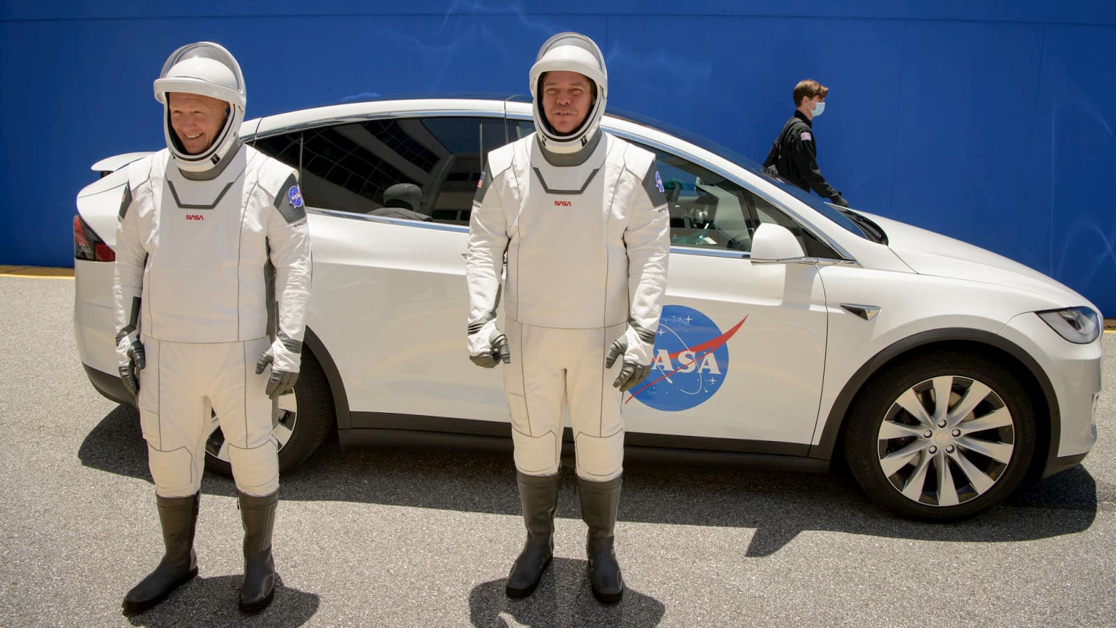 Os astronautas da NASA Douglas Hurley e Robert Behnken vestindo trajes espaciais SpaceX com um Tesla Model X atrás deles.