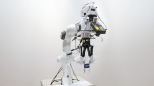 Robô com uma mão composta de duas placas segunrando uma garrafa de água de plástico. O robô também tem uma câmera apontada para sua mão.