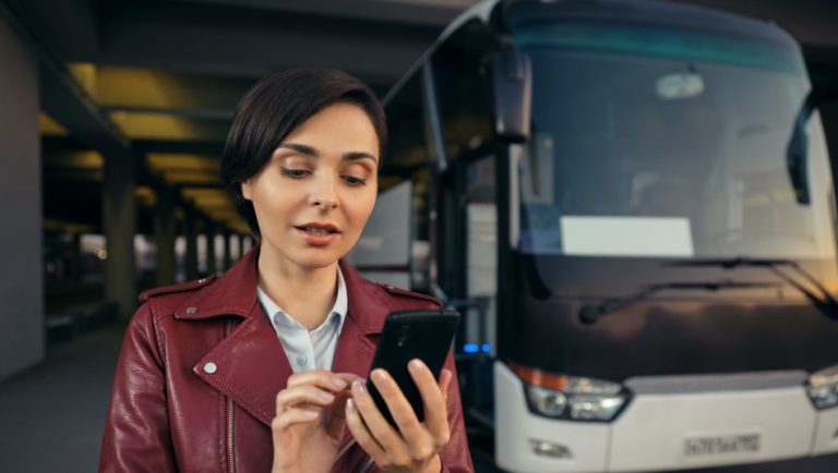 Imagem mostra mulher mexendo em um smartphone com um ônibus ao fundo. Crédito: BlaBlaCar